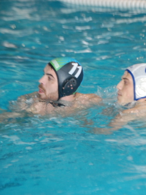 Dos jugadores de waterpolo en acción en una piscina azul, uno con un casco verde y el otro con un casco blanco y azul.