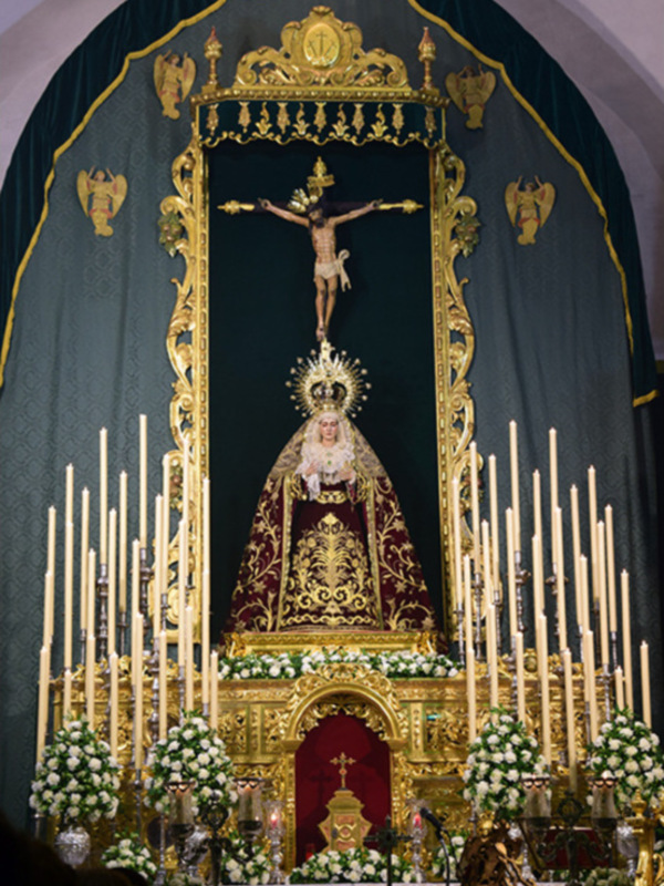 Altar con una imagen de la Virgen María y un crucifijo en el centro, rodeado de velas y flores blancas. La imagen está iluminada por luces doradas que resaltan su belleza y solemnidad.