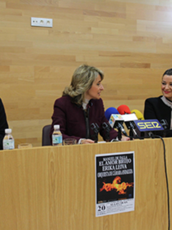 Tres personas en una mesa con micrófonos y carteles promocionales en un evento de prensa.