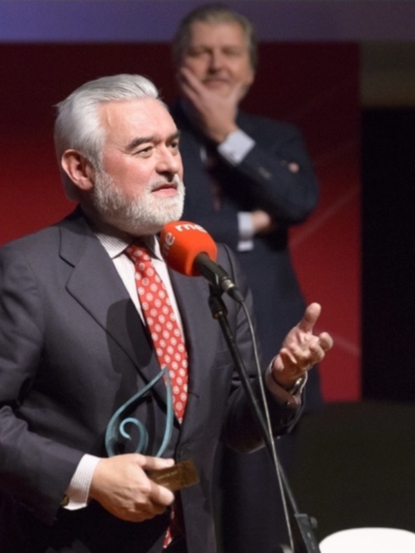 Hombre en traje negro y corbata con lunares rojos, sosteniendo un premio mientras habla en un micrófono. Fondo con elementos gráficos en rojo y negro.