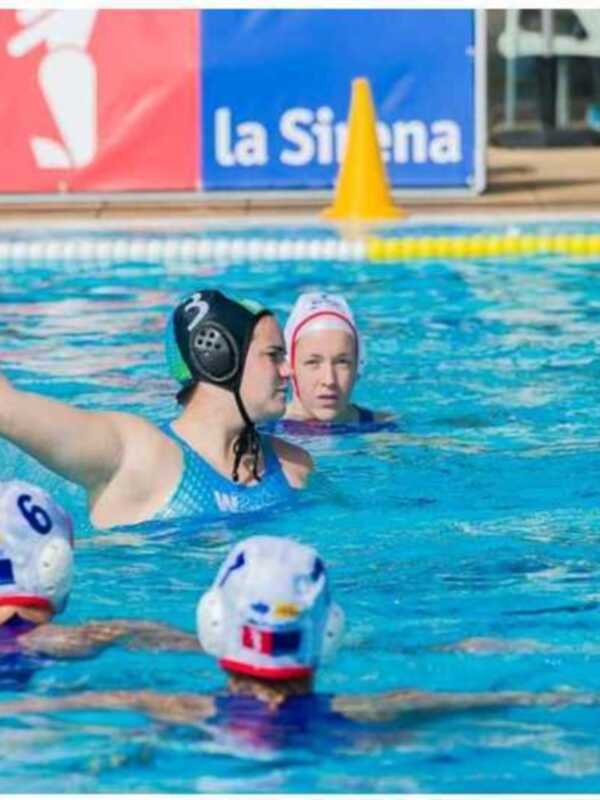 Jugadoras de waterpolo en acción durante un partido en una piscina de agua azul. Las jugadoras llevan cascos con banderas nacionales y están en el agua, con una bandera de la Sisueña en el fondo.