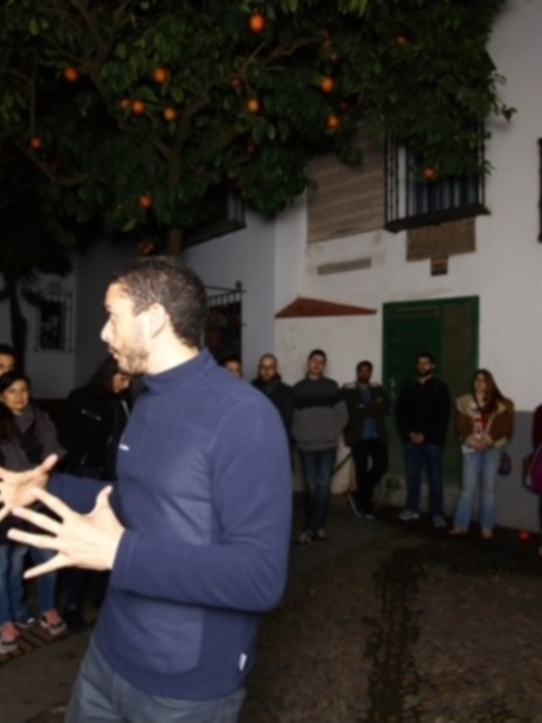 Una persona explica a un grupo de personas en una calle, con un árbol de naranjas en el fondo y una casa con el nombre 'Las Cadenas' en la esquina.