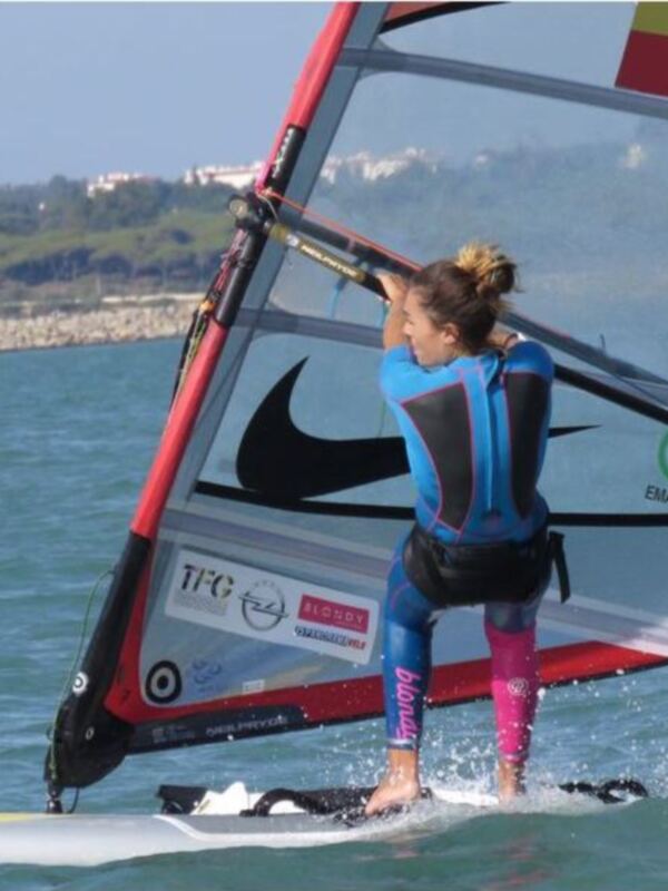 Una persona windsurfing en un agua azul con una tabla y un vela roja y negra. La persona está en el agua, balanceándose sobre la tabla mientras maneja la vela con una mano.