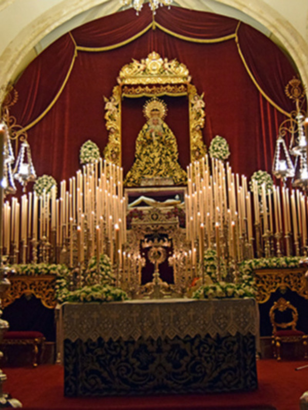 Interior de una iglesia con un altar adornado con velas y flores, rodeado de sillas y decoraciones doradas.