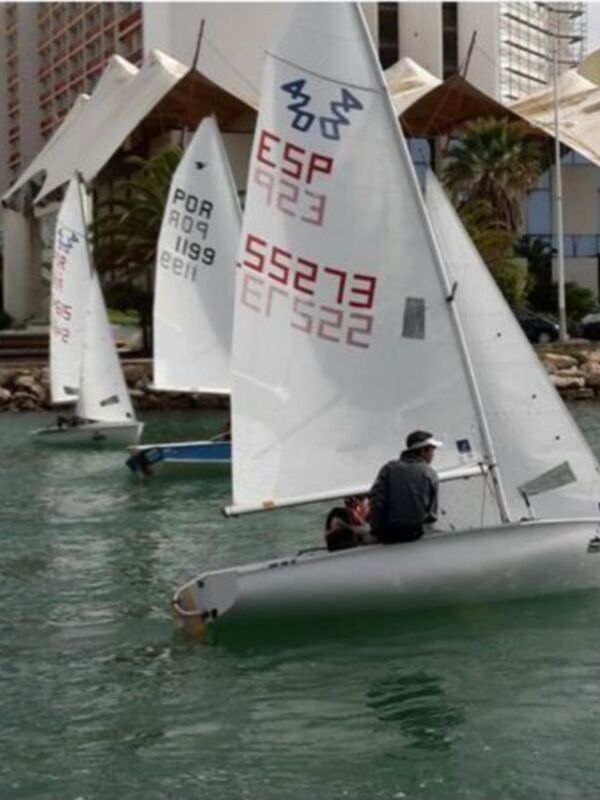 Competición de vela en el puerto con barcos navegando y espectadores en botes de resguardo.