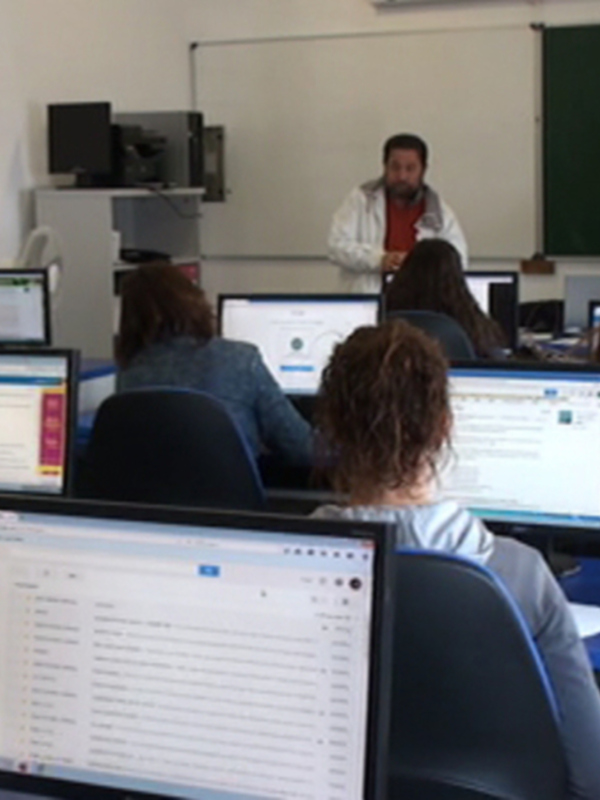 Clase de informática con estudiantes trabajando en computadoras y un profesor explicando en el pizarrón.