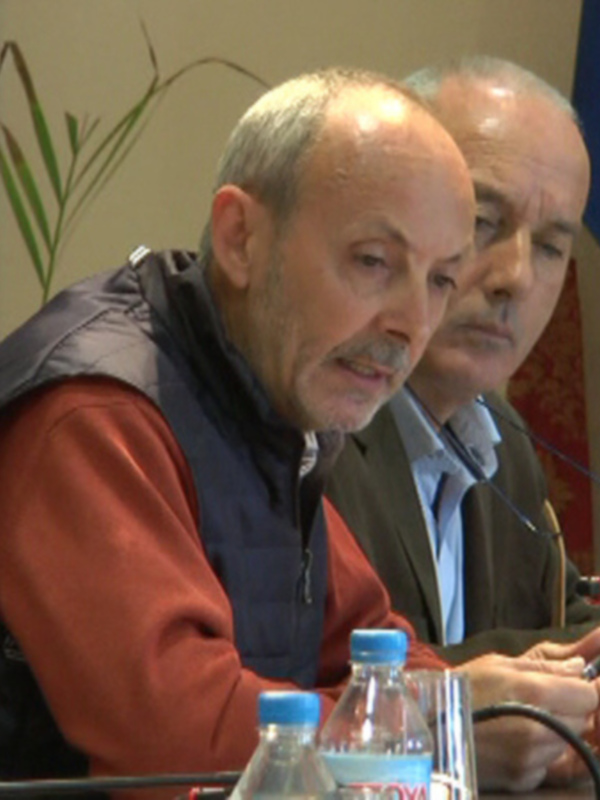 Conferencia de prensa con tres hombres sentados en una mesa con micrófonos y bandera de la UE en el fondo.