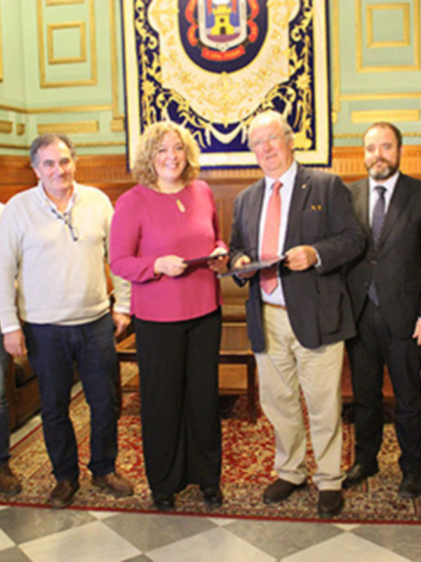 Gente en un evento formal, posando con un documento en una sala con decoración clásica y un escudo en el fondo.