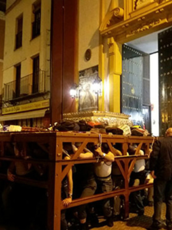 Fiesta religiosa con procesión en una calle de Sevilla, España, con personas portando un paso en un entorno urbano iluminado.