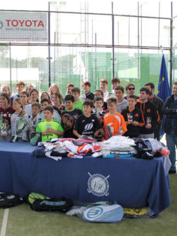 Celebración en un campo de pádel con jóvenes jugadores y autoridades, rodeados de material deportivo y banderas.