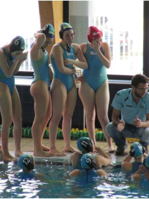 Niños y niñas practicando natación en una piscina interior, con un entrenador observando y guiando a los nadadores.