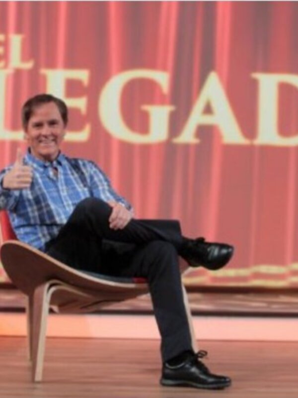 El hombre está sentado en un sillón de diseño moderno, con una sonrisa y un gesto de aprobación. La imagen se centra en él, destacando su expresión y la elegancia del sillón. El fondo es un escenario con una幕 de color rojo y dorado, que añade un ambiente formal a la escena.