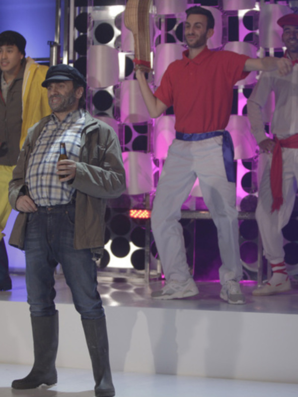 Un grupo de personas en una escena teatral, con un hombre en el centro vestido con una chaqueta y pantalones azules, sosteniendo un sombrero y una botella de cerveza, mientras otros personajes observan en el fondo.