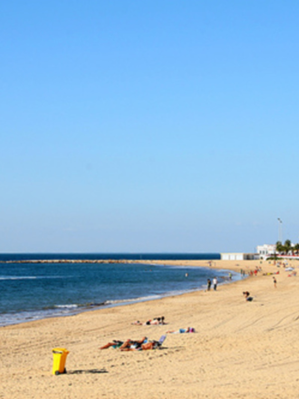 Paisaje de playa con faro y edificios en el horizonte bajo un cielo azul claro.