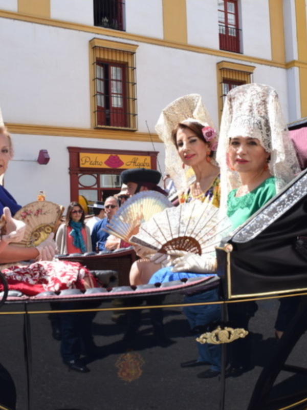 Una imagen de cuatro mujeres vestidas con trajes tradicionales españoles, posando en un carro negro. La escena parece ser parte de una celebración o evento cultural, con edificios blancos y amarillos al fondo.