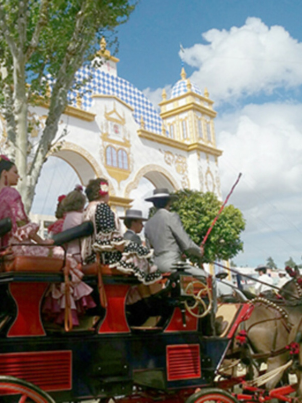 Un carro de caballos con decoraciones navideñas pasa frente a un edificio histórico con una torre de estilo gótico. El cielo está despejado con algunas nubes, y hay banderas ondeando en el fondo.