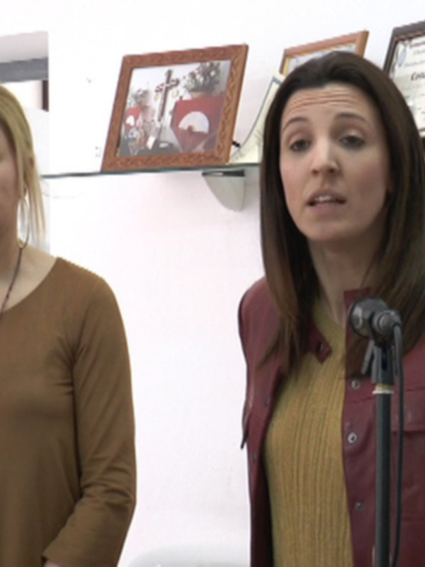 Dos mujeres hablando en un evento, una detrás de un micrófono, en un espacio con paredes blancas y marcos en la pared.