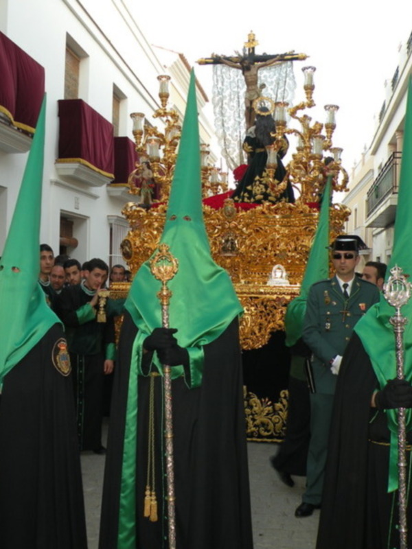 Procesión religiosa con hermandades en túnicas verdes y capas negras, portando un paso dorado con un crucificado, en una calle de edificios blancos.