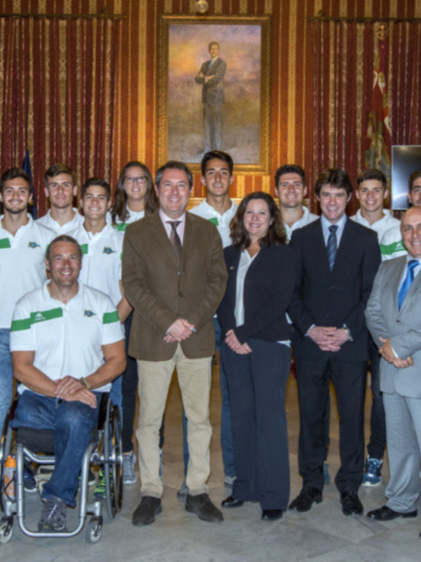 Equipo deportivo con autoridades en una ceremonia oficial en un salón decorado con cortinas rojas y retratos.