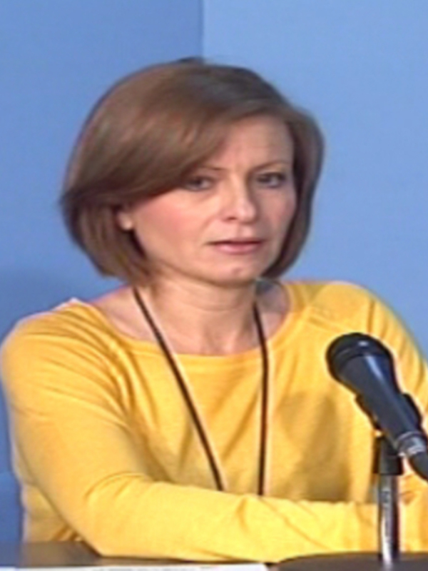 Una mujer con cabello corto y una camiseta amarilla se sienta frente a un micrófono, posiblemente en una conferencia o entrevista.