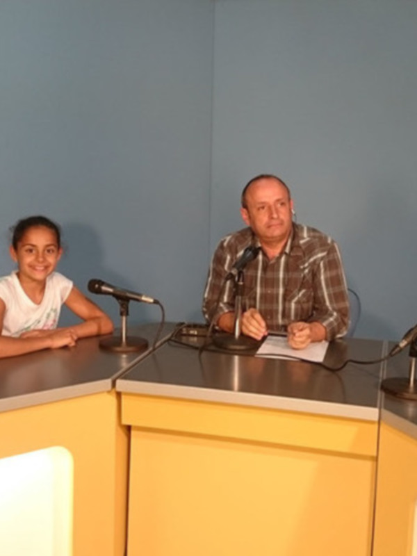 Una niña y un hombre sentados en un estudio de radio, con micrófonos y una mesa en el frente.
