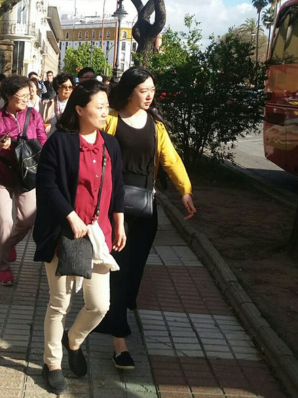 Dos mujeres caminan juntas por una calle adoquinada en un entorno urbano soleado, rodeadas de árboles y edificios.