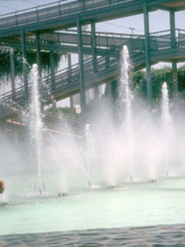 Fuente de agua en un parque con personas disfrutando y un puente en el fondo.
