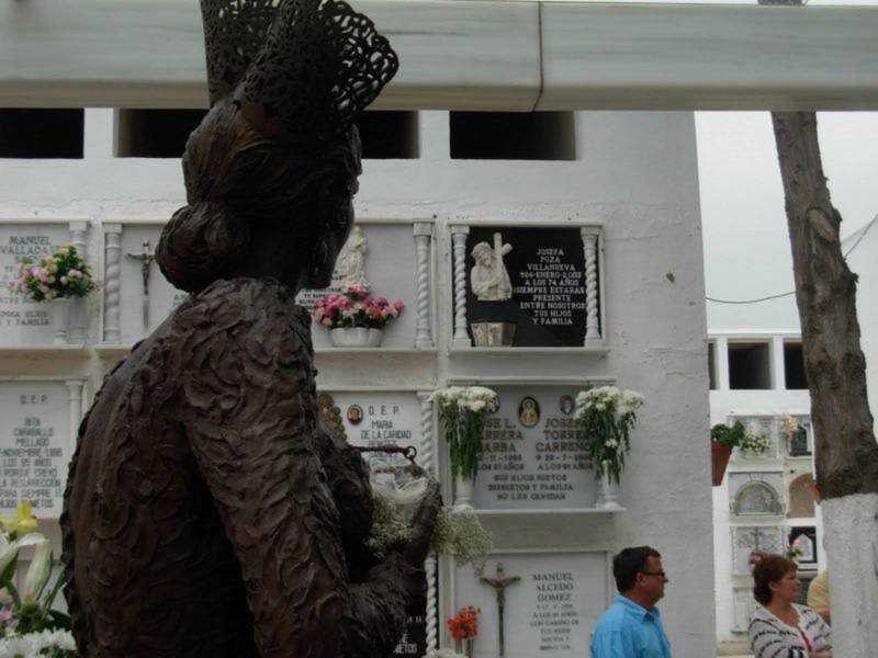 Un hombre camina junto a una estatua en un cementerio, rodeado de flores y placas con nombres de personas fallecidas.