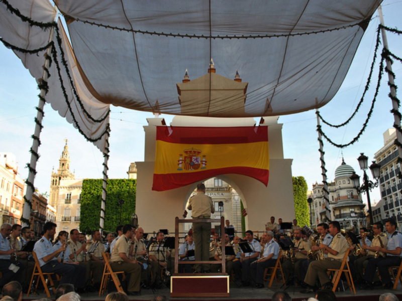 Concierto de banda militar en un escenario adornado con bandera española y decoración festiva, en un entorno urbano con edificios históricos al fondo.