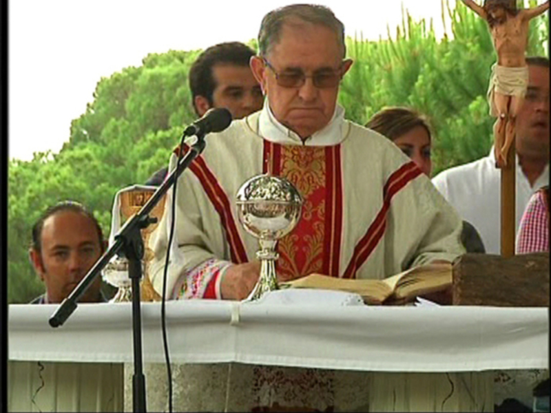 Un sacerdote en vestimenta religiosa oficia una ceremonia religiosa al aire libre, con un crucifijo en el fondo y un libro abierto en el altar.