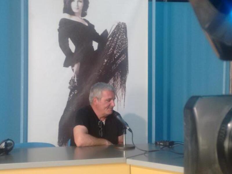 Persona hablando en un estudio de radio o televisión con un fondo de imagen de una mujer en traje negro.