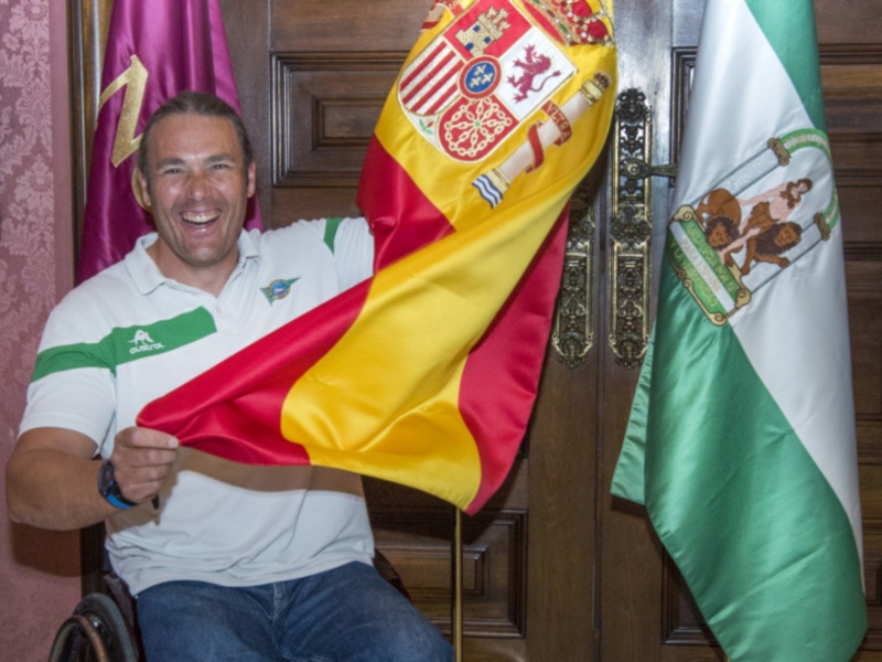Persona en silla de ruedas sonriendo con banderas española y andaluza al fondo.
