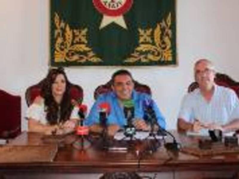 Conferencia de prensa con tres personas detrás de un escritorio con micros y bandera en el fondo.