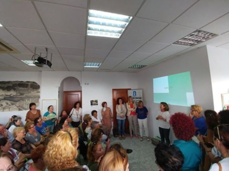 Una conferencia en un salón con una presentadora y un público interesado. La sala tiene una proyección en la pared y una puerta al fondo. La presentadora está en el centro, con el público alrededor. La iluminación es adecuada para el evento.