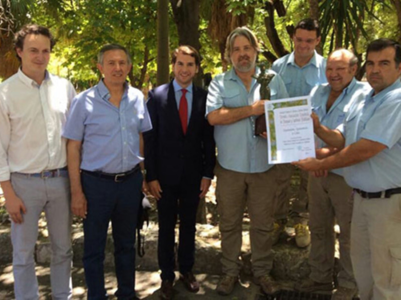 Grupo de personas celebrando la entrega de un reconocimiento, posando con un diploma en un entorno natural con árboles y vegetación.