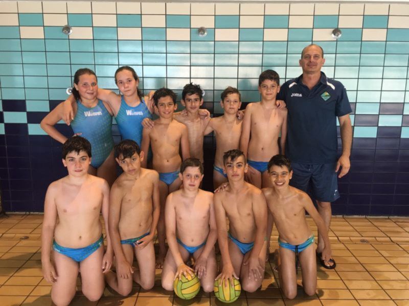 Equipo de natación infantil posando en la piscina, con dos balones de waterpolo en el suelo. El equipo está compuesto por niños y niñas, con un entrenador adulto a su lado. La piscina tiene azulejos de colores azul, blanco y turquesa.