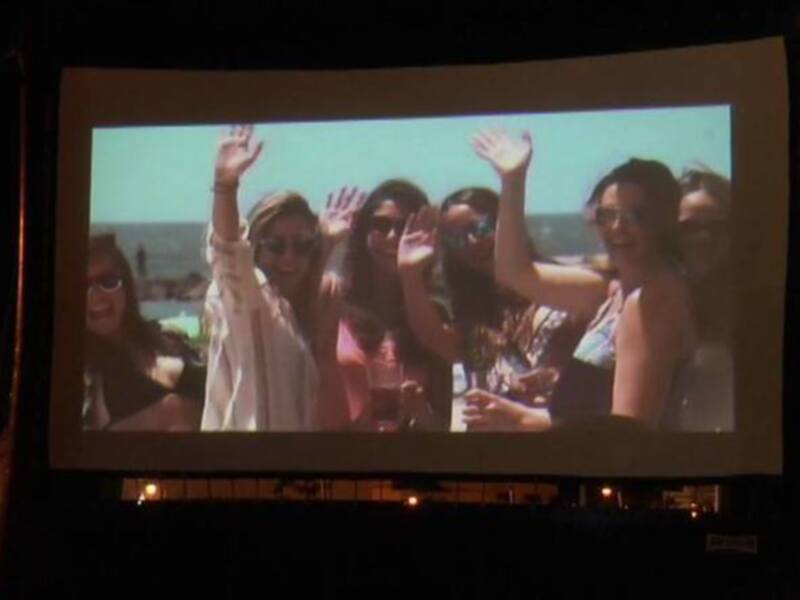 Amigas felices en la playa, celebrando juntas en una noche de verano. La imagen se proyecta en un gran pantalla al aire libre, iluminando su sonrisa y la brisa de la playa.