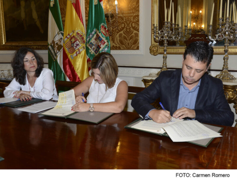 Firma de documentos en un entorno formal con banderas de España y Andalucía en el fondo.
