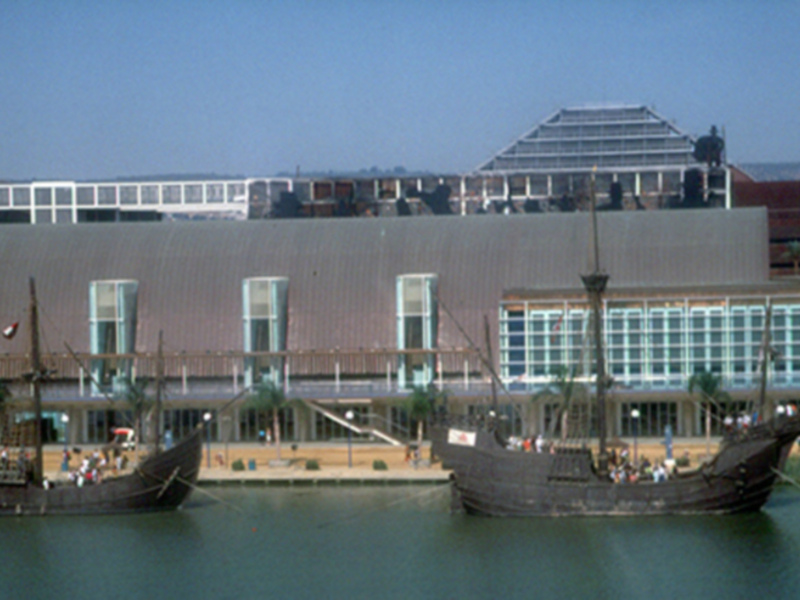 Barcos históricos flotando frente a un edificio moderno con una estructura curva y grandes ventanas. El cielo es azul claro y la agua es de color turquesa. El edificio parece ser un museo o centro cultural.