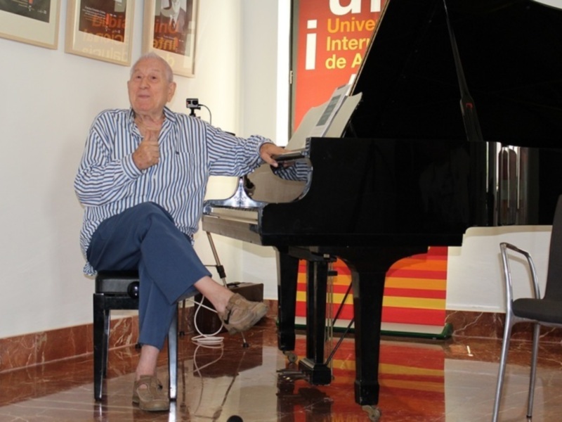Elderly man sitting on a chair next to a grand piano, appearing to be in a relaxed or contemplative pose.