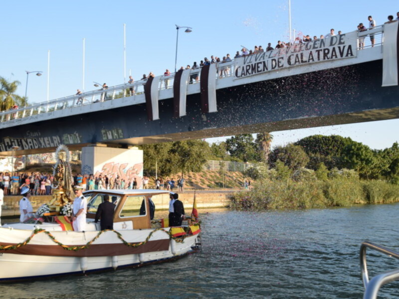 Celebración fluvial con barco adornado y confeti en el aire, bajo un puente con letreros de 'Viva la Virgen del Carmen de Calatrava'.