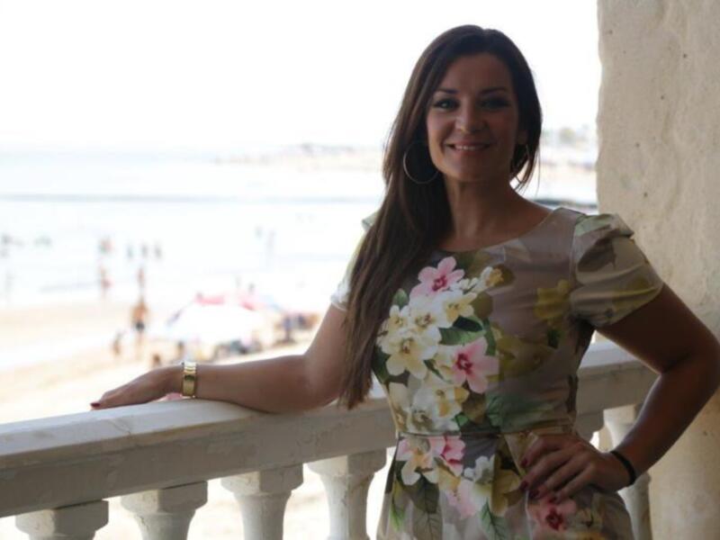 Una mujer con vestido floral posa en un balcón con vista al mar, iluminada por el sol. La imagen captura un momento relajado y soleado, con el mar y la playa en el fondo.