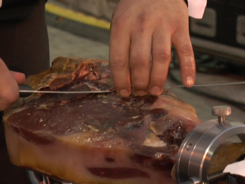 Un chef cortando un jamón con un cuchillo en una cocina profesional. El jamón parece estar bien asado y tiene una textura suave. La cocina tiene un ambiente de trabajo serio y profesional, con equipos de cocina en el fondo.