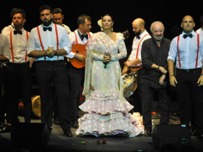 Una imagen de un grupo de personas en trajes formales, posiblemente una banda o coro, con un cantante en el centro vestido de flamenca. La escena parece ser parte de una actuación o concierto, con un ambiente festivo y celebrativo.