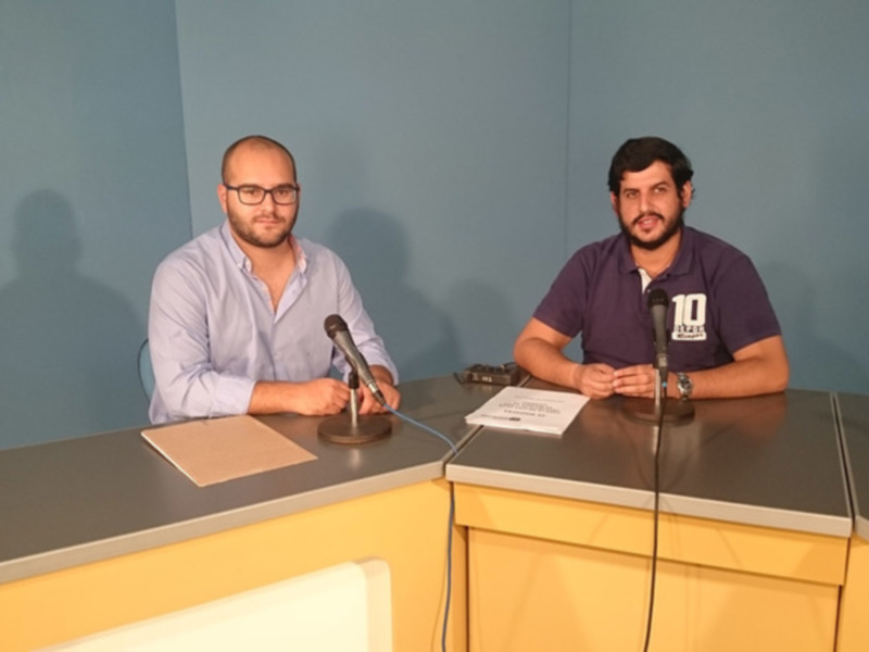Dos hombres sentados en una mesa con micrófonos, posiblemente en una entrevista o conferencia de prensa.