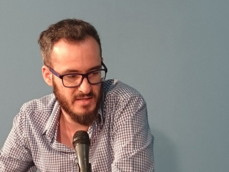 Hombre con gafas hablando en un evento con micrófono en la mesa.
