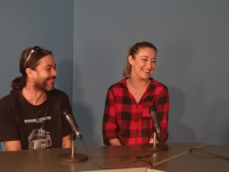 Dos personas sentadas en una mesa con micrófonos, posiblemente en una conferencia o presentación.