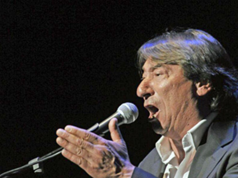 Un hombre con una camisa blanca y una chaqueta negra canta en un escenario con un micrófono en la mano. La iluminación es dramática, enfocándose en su cara y manos.