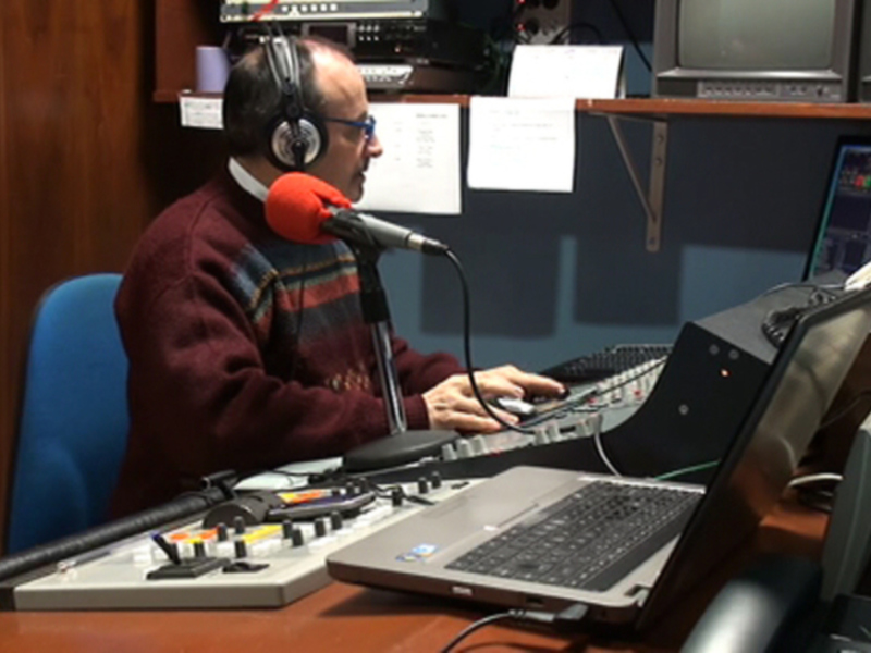 Persona en estudio de radio, transmitiendo con auriculares y micrófono, rodeada de equipos de audio y tecnología.