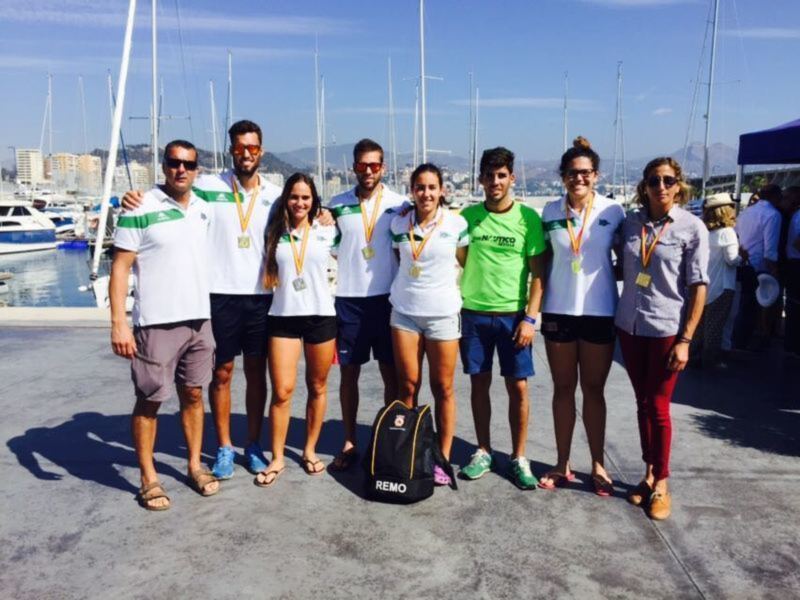 Un equipo de atletas posa con sus medallas de oro en un puerto deportivo, con un fondo de embarcaciones y un cielo azul claro.